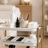 Salon trolley with lash tools and foaming cleanser beside a treatment bed in a warm neutral beauty studio.