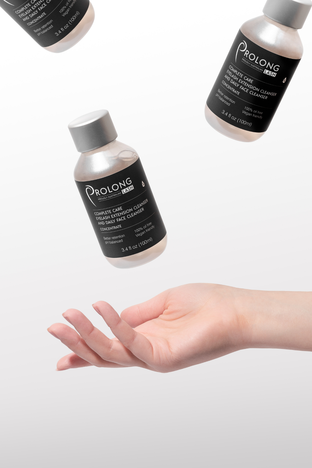 An outstretched hand hovering below three bottles of Prolong Lash Cleanser Concentrate which are floating above. The three bottles are small 100ml sizes plastic bottles with silver lids and black labels.